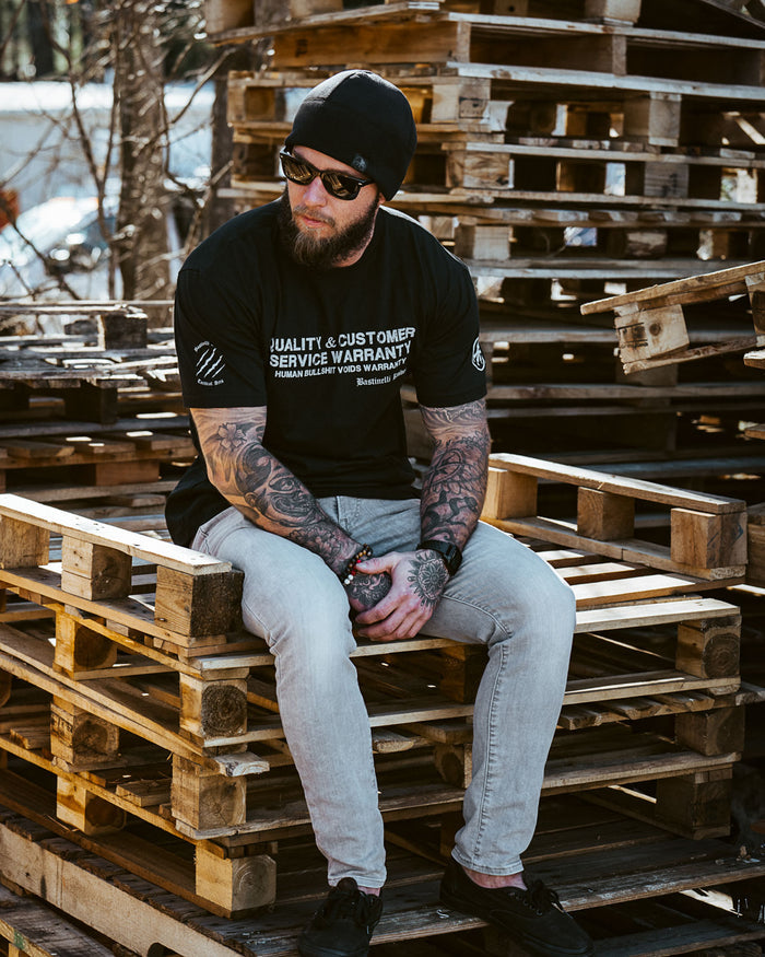 Tattooed man in black graphic t-shirt and beanie sitting on wooden pallets outdoors