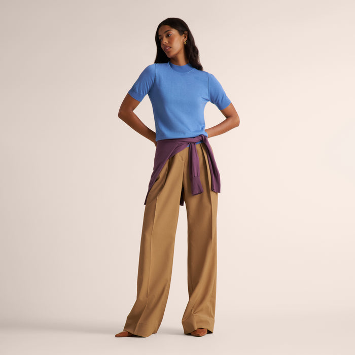 Woman wearing a mock neck short sleeve blue splendor sweater with wide leg beige pleated pants and purple sweater tied around her waist