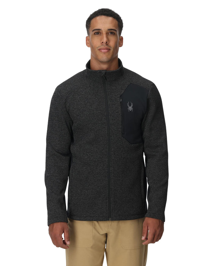 Man wearing a dark gray fleece jacket with a brand logo on a white background