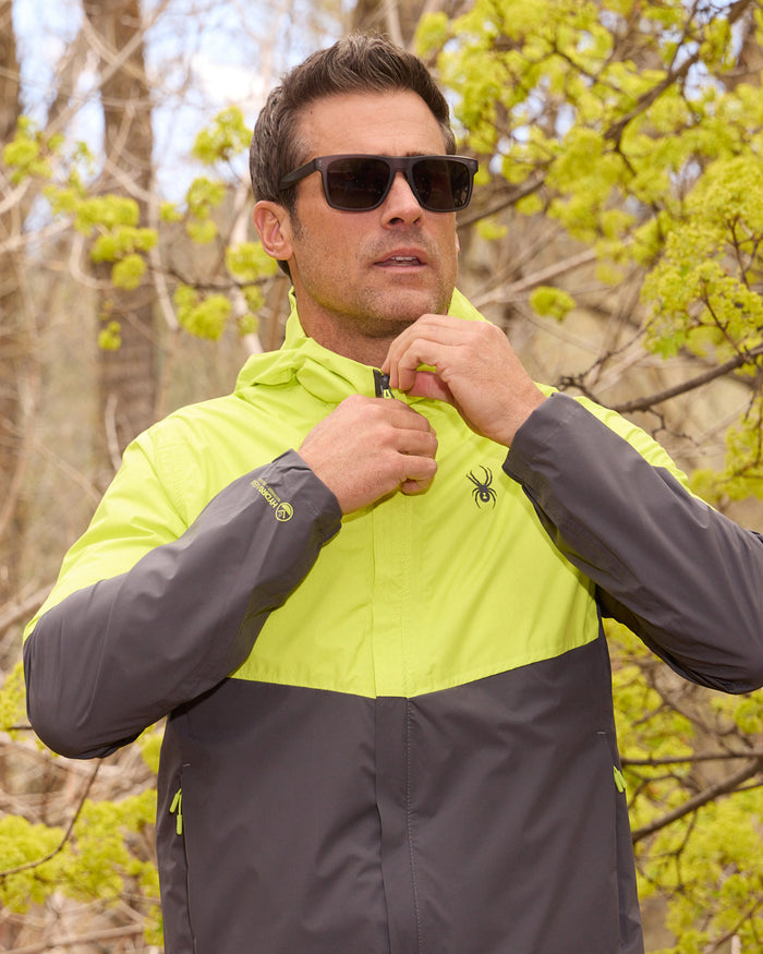 Man wearing a gray and yellow jacket with a brand logo, standing outdoors with trees in the background.
