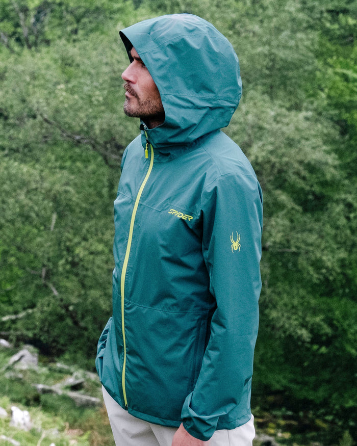 Person wearing a teal jacket with a brand logo in a forest setting