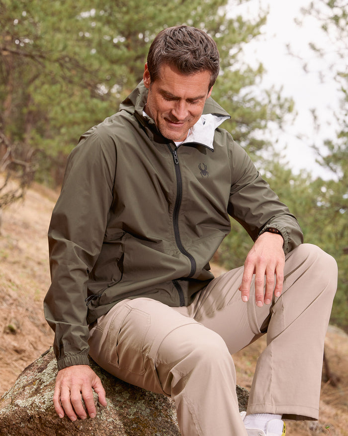 Man sitting on a rock in a forest wearing a green jacket and beige pants.