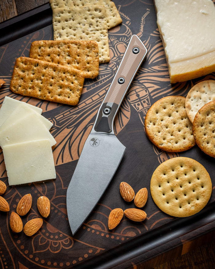 Microtech Culinary Santoku 4.2" Knife with coyote tan G10 handle, titanium bolsters and stonewashed stainless steel blade on a dragon fly cutting board surrounded by cheeses, crackers and nuts. Premium professional kitchen knife for precision chopping and modern cooking.
