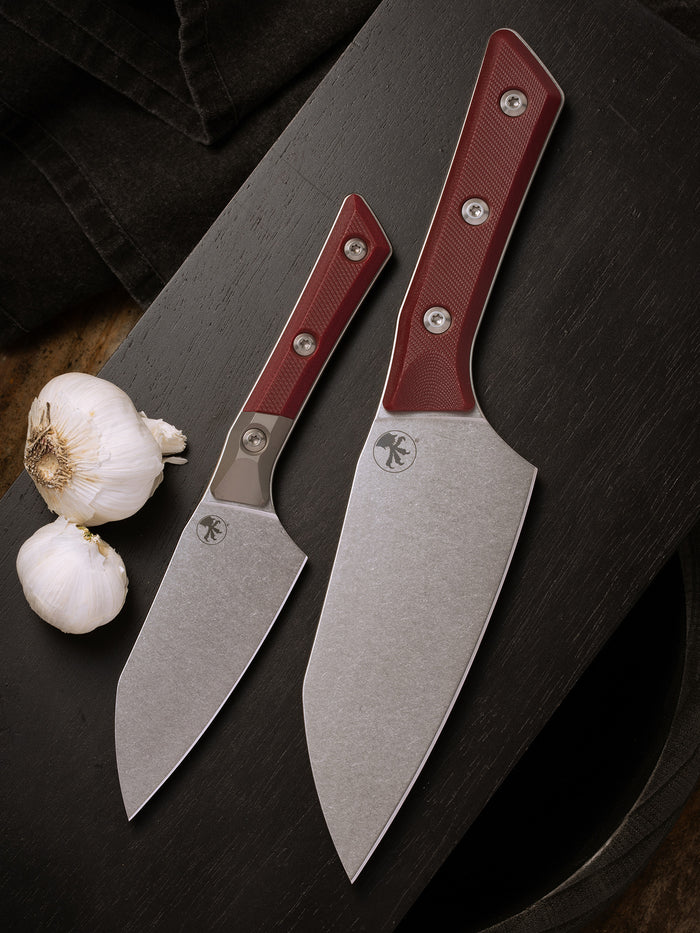 Microtech Culinary Santoku 4.2" and 6" Knife with merlot G10 handle, titanium bolsters (on the 4.2") and stonewashed stainless steel blade on a black cutting board surrounded by garlic. Premium professional kitchen knife for precision chopping and modern cooking.